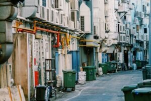 A narrow urban alley lined with numerous air conditioning units and trash bins.